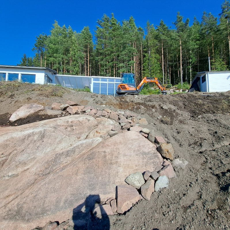 Minigraver foran en lav bolig under utgraving, med fjell og jordmasser i forgrunnen